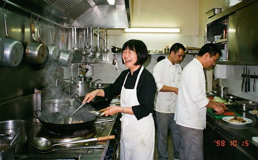 Una vecchia foto a pellicola del 1998 mostra Cinzia, che ride mentre cucina con un wok fumante in una cucina professionale, indossando una divisa nera da cuoca e un grembiule bianco.